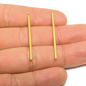 Puede incluir: Dos componentes de pendientes dorados, de forma rectangular. Cada barra tiene un pequeño agujero en la parte superior. Los pendientes se sostienen en la mano de una persona sobre un fondo blanco. Suministros para la fabricación de joyas.