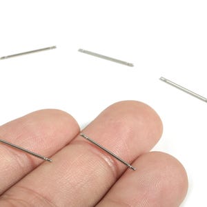 Peut inclure: Clous d'oreilles argentés avec un design de barre droite. Plusieurs clous sont dispersés sur un fond blanc, dont deux sont tenus entre les doigts. Les clous ont de petites extrémités arrondies.