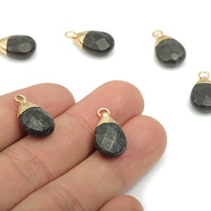 Puede incluir: Una colección de colgantes de piedras preciosas en forma de lágrima. Cada colgante es una piedra gris oscura facetada con una tapa dorada y un pequeño lazo para sujetarlo a una cadena u otro componente de joyería. Los colgantes se muestran sobre un fondo blanco.