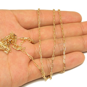 Op de afbeelding: Een goudkleurige ketting met een delicaat, rechthoekig schakelontwerp. De ketting wordt op een hand getoond, waarbij de lengte en stijl worden getoond. De ketting heeft een heldere, glanzende goudkleur.