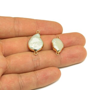 Puede incluir: Dos colgantes de perlas blancas irregulares con bordes y bucles de metal dorado. Los colgantes se sostienen en una mano sobre un fondo blanco. Las perlas tienen una superficie texturizada.