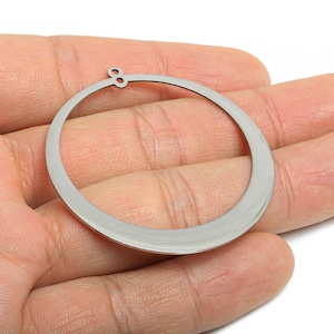 Peut inclure: Un composant de boucle d'oreille circulaire de couleur argentée avec une petite boucle en haut. Le composant de boucle d'oreille est tenu dans la main d'une personne, et le fond est blanc.