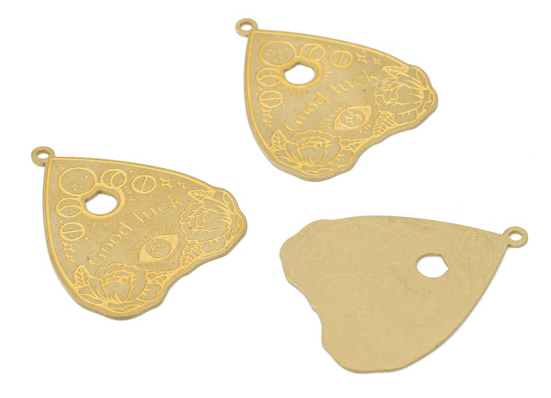 May include: Three gold-colored metal charms in the shape of a triangle with rounded edges. The charms have a hole at the top for hanging. Two of the charms have an etched design of a stylized eye, flowers, and the words "Good Luck". The third charm is plain.