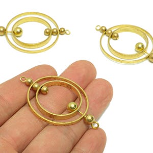 May include: Gold-coloured metal jewellery components. The components feature concentric circles and small spheres. The design includes a central ring with two smaller rings inside, and small spheres attached to the outer rings. The components have a textured finish.