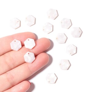 May include: A collection of white, hexagonal beads with a pearlescent finish. Some beads are held in a hand, while others are scattered on a white surface. Each bead has a small hole for stringing.