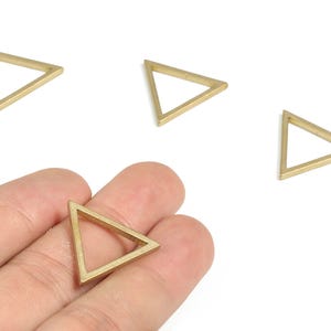 以下が含まれることがあります： 金色の三角形のジュエリーパーツが4つ。1つは手に持たれ、他は白い表面に散らばっています。三角形は中空で、細いフレームで形が縁取られています。 ジュエリー作りに適しています。
