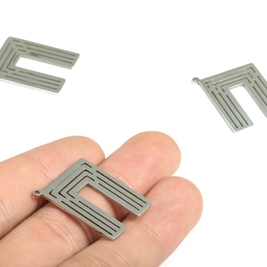 Puede incluir: Tres colgantes de metal plateado con un diseño geométrico en forma de U. Cada colgante tiene un pequeño agujero para ensartar y múltiples líneas paralelas dentro de la forma de U. Un colgante se sostiene en una mano.