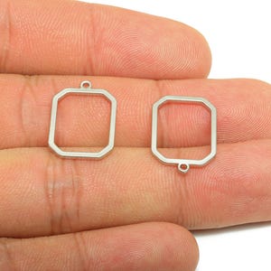 May include: Two silver-colored, open-frame geometric charms. Each charm has a rectangular shape with beveled edges and a small loop for attachment. The charms are held in a hand against a white background.