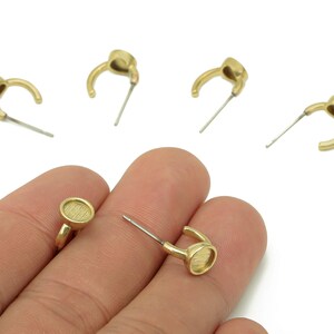 May include: Gold-coloured earring components. The earrings feature a curved design with a small, round, textured setting for embellishments. The posts are silver. The earrings are displayed against a white background.