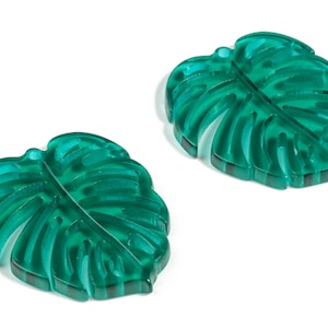 May include: Two teal green monstera leaf shaped acrylic charms. The leaves have a raised, textured design.