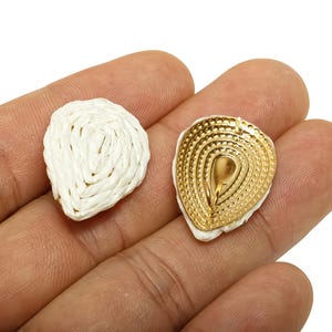 May include: Two teardrop-shaped earrings. One is white woven material, the other is gold-coloured metal with a textured design. The earrings are displayed on a hand against a white background. Jewellery.