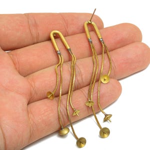 Peut inclure: Boucles d'oreilles pendantes dorées au design unique. Chaque boucle d'oreille présente un dessus incurvé relié à plusieurs chaînes délicates. De petits accents ronds dorés sont espacés le long des chaînes, créant un look fluide et élégant. Les boucles d'oreilles sont présentées sur fond neutre.