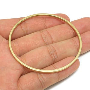 May include: A gold-coloured, circular bangle bracelet. The bracelet has a simple, minimalist design with a smooth, polished surface. The bangle is held in a hand, showcasing its size and shape. The jewellery is likely made of metal.