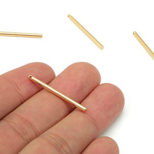 May include: Close-up of several gold-coloured, straight bar-shaped jewellery findings. One bar is held in a hand, with others scattered around. Each bar has a small hole at one end, and rounded tips.