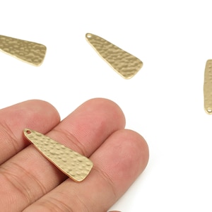 May include: Gold-colored metal pendants with a hammered texture. The pendants are elongated with a rounded end and a small hole for stringing. One pendant is held in a hand.