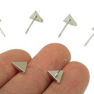 May include: Silver-colored triangle stud earrings. The earrings have a simple, geometric design with a triangular shape. The image shows several pairs of the earrings, with some displayed on a person's hand.
