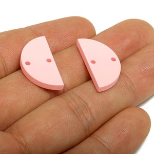 以下が含まれることがあります： 2つの淡いピンク色の半円形のクラフトコンポーネント。それぞれに2つの小さな穴があります。コンポーネントは手で持たれており、ジュエリー作りやその他のクラフトプロジェクトに使用される可能性があります。