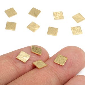 Puede incluir: Pequeños dijes cuadrados de metal dorado con superficie texturizada. Varios dijes están dispersos sobre un fondo blanco, con algunos sostenidos entre los dedos. Estos dijes son ideales para la fabricación de joyas.