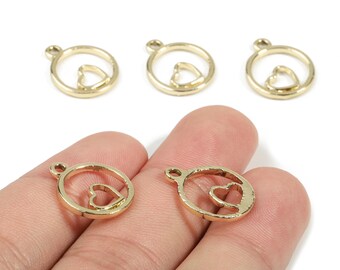 May include: Gold-colored metal charms with a heart-shaped cutout inside a circular frame. The charms have a small loop at the top for attaching to jewelry or other crafts. Several charms are visible, some held in a hand.