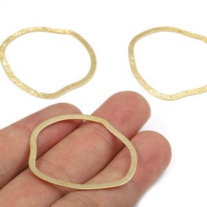 May include: Three gold-coloured, irregular-shaped metal jewellery components. One is held in a hand, while two others are visible in the background. The components have a textured surface and a wavy, organic design, suitable for jewellery making.