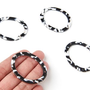 Peut inclure: Quatre composants de bijoux ovales avec un motif tacheté noir et blanc. L'un est tenu par une main. Chaque composant comporte deux petits trous. Ces composants conviennent à la fabrication de bijoux.