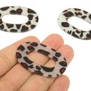 May include: Three oval-shaped leopard print acrylic charms. The charms have a clear background with black and brown spots. One charm is held in a hand. These charms are suitable for jewellery making and crafting.