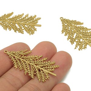 May include: Gold-coloured metal fern leaf charms. The charms have a detailed, openwork design, with a small loop at the top for attaching to jewellery or other crafts. The image shows three charms, one held in a hand.