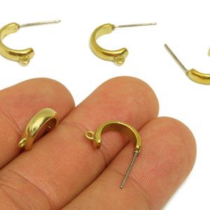Könnte beinhalten: Gezeigt werden mehrere goldfarbene Creolen-Ohrringkomponenten. Die Ohrringe haben ein gebogenes, halbkreisförmiges Design mit einem Stecker zum Durchstechen und einer kleinen Schlaufe zum Anbringen zusätzlicher Elemente. Die Komponenten sind vor weißem Hintergrund.