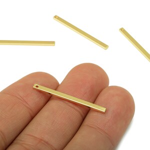 May include: Gold-colored rectangular bar charms with a small hole at one end. Several charms are scattered on a white background, with one held in a hand. These charms are likely used for jewelry making or crafting.