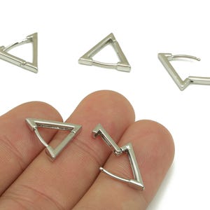 May include: Silver-toned triangular hoop earrings. The earrings have a geometric design with a hinged closure. Several earrings are displayed, some open and some closed, against a white background.