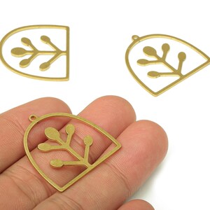 May include: Three gold-colored metal charms with a botanical design. The charms feature a stylized plant with rounded leaves within a teardrop or arched frame. One charm is held in a hand.