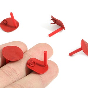 May include: Red, leaf-shaped stud earrings with a post and a small loop detail. The earrings are shown in a close-up shot, with some pieces scattered around. The earrings are made of a solid material.