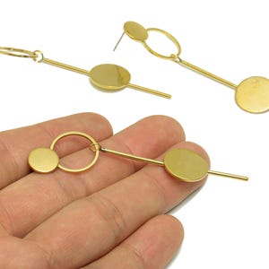 Pode incluir: Brincos geométricos dourados com um pino circular, um círculo pequeno e um disco maior conectados por barras retas. Os brincos têm fecho de pino e um design moderno e minimalista.
