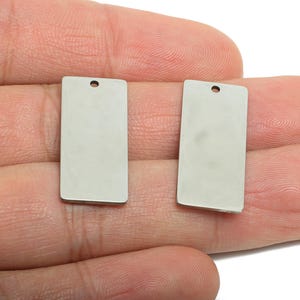 Puede incluir: Dos colgantes rectangulares de metal plateado con un pequeño agujero en la parte superior para ensartar. Los colgantes se sostienen en una mano sobre un fondo blanco. Los colgantes son probablemente para la fabricación de joyas.