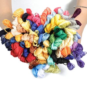 Puede incluir: Una vibrante colección de hilos de bordar en una variedad de colores, incluyendo rojo, amarillo, azul, verde y morado. Los hilos están agrupados, creando una exhibición colorida. La imagen está sobre un fondo blanco.