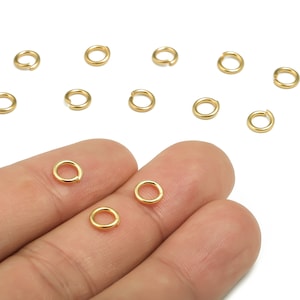 Op de afbeelding: Een close-up van een hand die verschillende gouden springringen vasthoudt. De springringen zijn klein en rond en perfect voor het maken van sieraden.