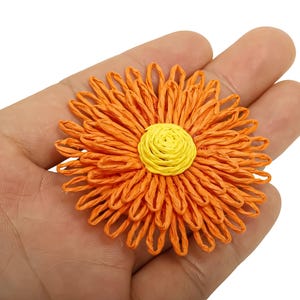 May include: Close-up of a handcrafted orange and yellow raffia flower. The flower has a bright orange outer layer with a yellow center. The raffia is woven to create a detailed floral design.