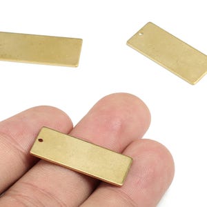 May include: Three rectangular brass metal charms with a small hole at the top for jewelry making. One charm is held in a hand. The charms are a warm gold color and have a smooth, matte finish. They are blank and ready for customization.