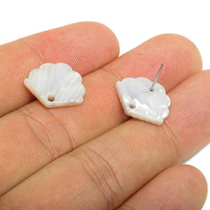 May include: Two white seashell-shaped stud earrings. One has a post for piercing, the other a small hole. The earrings are held in a hand, showing their pearly, iridescent finish. Ideal for beach-themed jewellery.