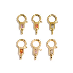 May include: Six gold-toned lobster claw clasps arranged in two rows. Each clasp features a circular ring at the top, a rectangular body with a small embedded baguette-cut gemstone in various colours including pink, orange, and clear, and a small loop at the base.