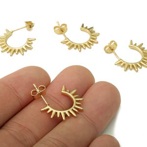 May include: Gold-colored sunburst hoop earrings. The earrings have a half-circle design with pointed rays extending outwards. The image shows several pairs of earrings, with one held in a hand. The earrings are on a white background.