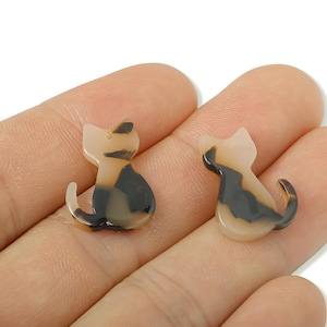 May include: Two small cat-shaped charms. The charms are a mix of tan, brown, and black, with a glossy finish. The cat charms are sitting upright with their tails curled. These charms could be used for jewelry making or other crafts.