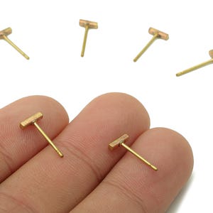 May include: A collection of small, gold-coloured bar stud earrings against a white backdrop. Two earrings are held between fingers, with others scattered around. The earrings feature a simple, minimalist design.