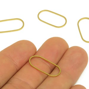 May include: Several small, gold-colored oval-shaped metal jewelry findings. One is held in a hand, with others scattered nearby. The findings have a simple, open design, suitable for jewelry making or crafting projects.