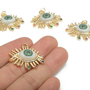 May include: Gold-colored eye-shaped pendants with a green iris and white background. The pendants have a sunburst design with colorful gemstones. The eye pendants are shown on a white background.