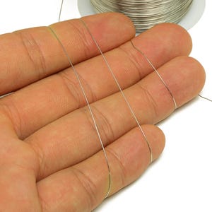 Può includere: Primo piano di sottili fili di filo argentato tenuti tra le dita. Una bobina dello stesso filo è visibile sullo sfondo. Il filo sembra essere utilizzato per la creazione di gioielli o artigianato.