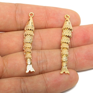 May include: Two gold-coloured fish-shaped pendants. Each pendant has a loop at the top for hanging, a textured body, and a detailed tail fin. The pendants are held in a hand against a white background.