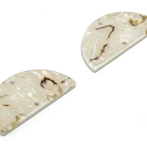 Może przedstawiać: Dwa perłowe, półksiężycowate elementy rzemieślnicze. Każdy ma kremowobiałą podstawę z brązowymi i fioletowymi żyłkami. Mają błyszczące wykończenie i trzy małe otwory do nawlekania lub mocowania. Prawdopodobnie są używane do tworzenia biżuterii lub innych prac rzemieślniczych.