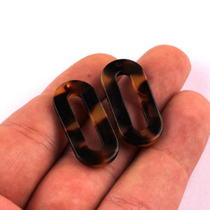 May include: Two oval-shaped, tortoise shell pattern pendants. Each pendant has a hole at the top for stringing. The pendants are held in a person's hand against a white background. The pendants are made of a glossy material.