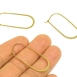 May include: Gold-colored oval hoop earrings. The earrings have a simple, elongated oval shape with a hook closure. The image shows three earrings, with one held in a hand. The background is white.